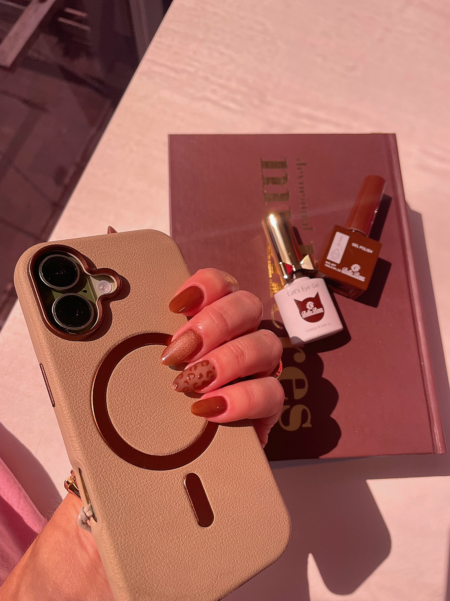 Mão com unhas em tom terracota segurando o celular ao lado dos esmaltes